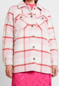 Vaate vaaleanpunaisella ja valkoisella ruudullisella kuviolla, jossa on pehmeä, teksturoitu kangas. Tässä takissa on kaksi rintataskua ja puunapit. Korostettuna punaisella ruudukko-kuviolla.