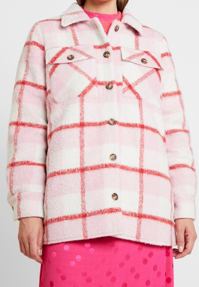 Vaate vaaleanpunaisella ja valkoisella ruudullisella kuviolla, jossa on pehmeä, teksturoitu kangas. Tässä takissa on kaksi rintataskua ja puunapit. Korostettuna punaisella ruudukko-kuviolla.