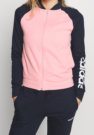 Kobieta ubrana w różowo-granatową kurtkę na zamek z logo "adidas" na rękawie oraz granatowe spodnie dresowe z małym logo adidas przy kieszeni.