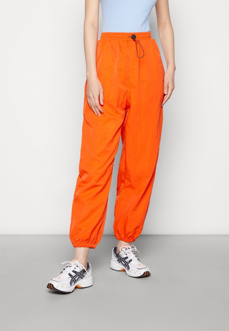 Personne portant un pantalon ample orange vif avec des chevilles élastiques, un haut bleu clair et des chaussures de sport blanches avec des détails noirs et orange.