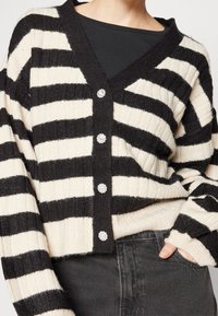 Czarny i kremowy kardigan w paski z dekoltem w serek, o prążkowanej fakturze, z czterema ozdobnymi guzikami. Elastyczne mankiety i dół. Nosi się go na czarnej bluzce.