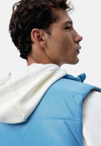 Veste doudoune bleu clair avec un hoodie blanc drapé sur l'épaule. Texture lisse, fermeture éclair à l'avant et détail de col haut.