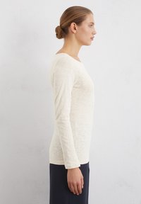 Langärmliges, cremefarbenes Oberteil aus strukturiertem Stoff mit Rundhalsausschnitt, figurbetonter Silhouette und geradem Saum, kombiniert mit dunklen Hosen.