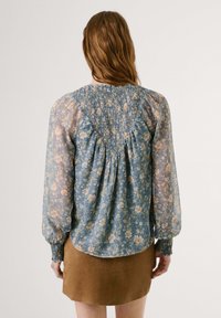 Blusa floreale con tessuto blu semitrasparente, schiena arricciata, maniche voluminose e dettaglio arricciato all'orlo. Abbinata a una gonna marrone.