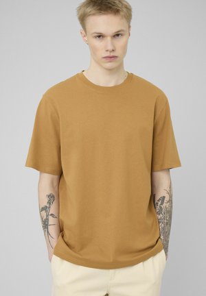 Jeune homme blond avec des tatouages sur les deux avant-bras, portant un t-shirt manches courtes moutarde et un pantalon beige clair sur un fond gris uni.