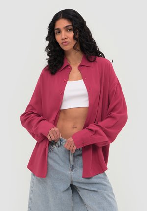 Frau mit lockigem dunklem Haar trägt ein übergroßes dunkelrosa Hemd über einem weißen Croptop und hellblaue High-Waist-Jeans, steht vor einem schlichten Hintergrund.