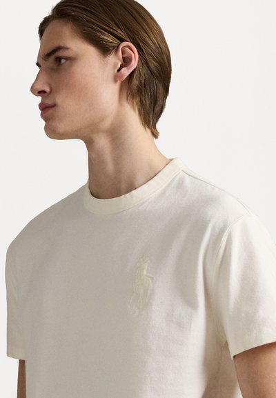 Camiseta de algodón color crema con cuello redondo, que presenta un logotipo bordado tonal en el pecho y mangas cortas. Textura suave.