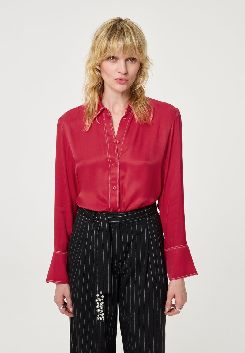Rode zijden blouse met een knoopsluiting aan de voorkant, puntige kraag en klokmouwen; gecombineerd met zwarte broek met streepjes en decoratieve hardware.