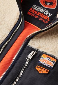 Fodrad jacka med fleecefoder i marinblå yttertyg, orangea meshdetaljer och silvriga dragkedjor. Har bergsinspirerade broderade märken.