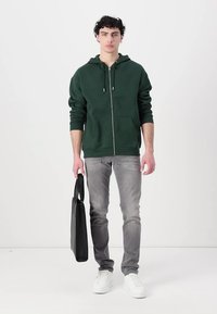 Haut à zip de couleur vert foncé, jeans gris, baskets blanches, tenant un sac fourre-tout noir. Design simple avec cordons de serrage et poches avant.