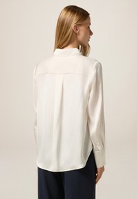 Blusa di seta color crema con maniche lunghe, una piega sul retro e un colletto standard. Presenta polsini con bottoni e un orlo curvo nella parte inferiore.