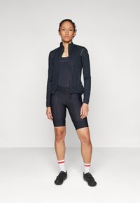 Black cycling jacket with a zip front, fitted black bib shorts, and black shoes. Accented with red and white striped socks. Smooth textures.
