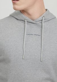 Szürke kapucnis sweatshirt zsinórokkal, kicsi tengerészkék nyomattal, amelyen a "CASUAL FRIDAY" felirat olvasható. Puha, texturált anyag, alapvető dizájn.