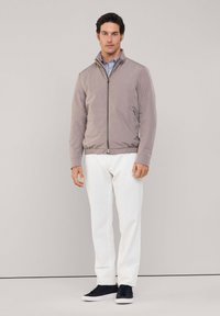Giacca bomber grigio chiaro con zip frontale, tasche laterali e polsini a coste, abbinata a pantaloni bianchi e sneakers scure.