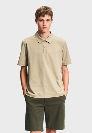 Jeune homme aux cheveux courts portant un polo beige et un short vert foncé, debout les mains dans les poches devant un fond uni.