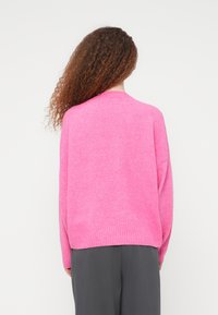 Γυναίκα με μακριά σγουρά μαλλιά, φορώντας ένα χαλαρό, φωτεινό ροζ πουλόβερ και σκούρο γκρι παντελόνι, με την πλάτη στραμμένη προς το άσπρο απλό φόντο.