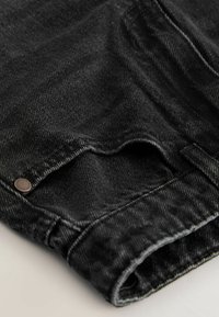 Gros plan d’un jean en denim noir plié montrant la texture du tissu, les détails des coutures et un rivet métallique dans le coin de la poche.