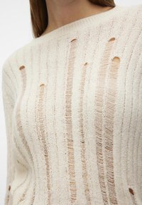 Pull en tricot couleur crème, avec des rayures verticales et des trous irréguliers partout, un grand col et une texture douce.