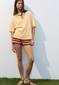 T-shirt surdimensionné rayé jaune et blanc, associé à un short en crochet multicolore avec des rayures horizontales. Sandales marron sur l'herbe.