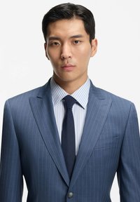 Niebieski garnitur w cienkie paski z klapą z nacięciem, zestawiony z jasnoniebieską i białą koszulą w paski oraz ciemnym krawatem. Gładka tkanina, dopasowany krój.