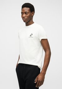 Νεαρός άνδρας που φοράει λευκό μπλουζάκι με μικρό λογότυπο φοίνικα και μαύρο παντελόνι, στέκεται μπροστά σε απλό φωτεινό φόντο.