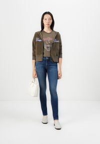 Maslinasta velura zip-up dukserica s izvezenim tekstom, grafička majica, plave uske traperice, bijela torba i svijetle cipele na slip-on.