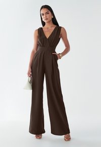 Smeđa kombinezon bez rukava s V-izrezom, strukom naglašenim remenom, hlačama širokih nogavica i bočnim džepovima. Glatki materijal s elegantnim, modernim dizajnom.