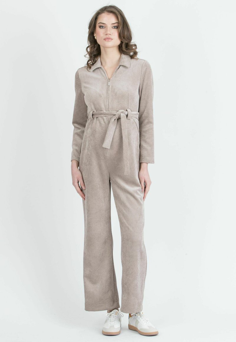 Salopette in corduroy beige con maniche lunghe, pantaloni lunghi, zip frontale e cintura annodata in vita. Il modello indossa sneakers bianche.