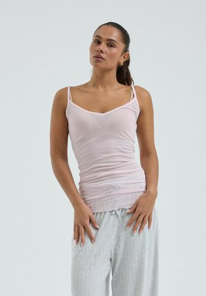 Femme aux cheveux noirs attachés en queue de cheval, portant un débardeur rose clair et un pantalon ample gris clair, debout devant un fond uni.