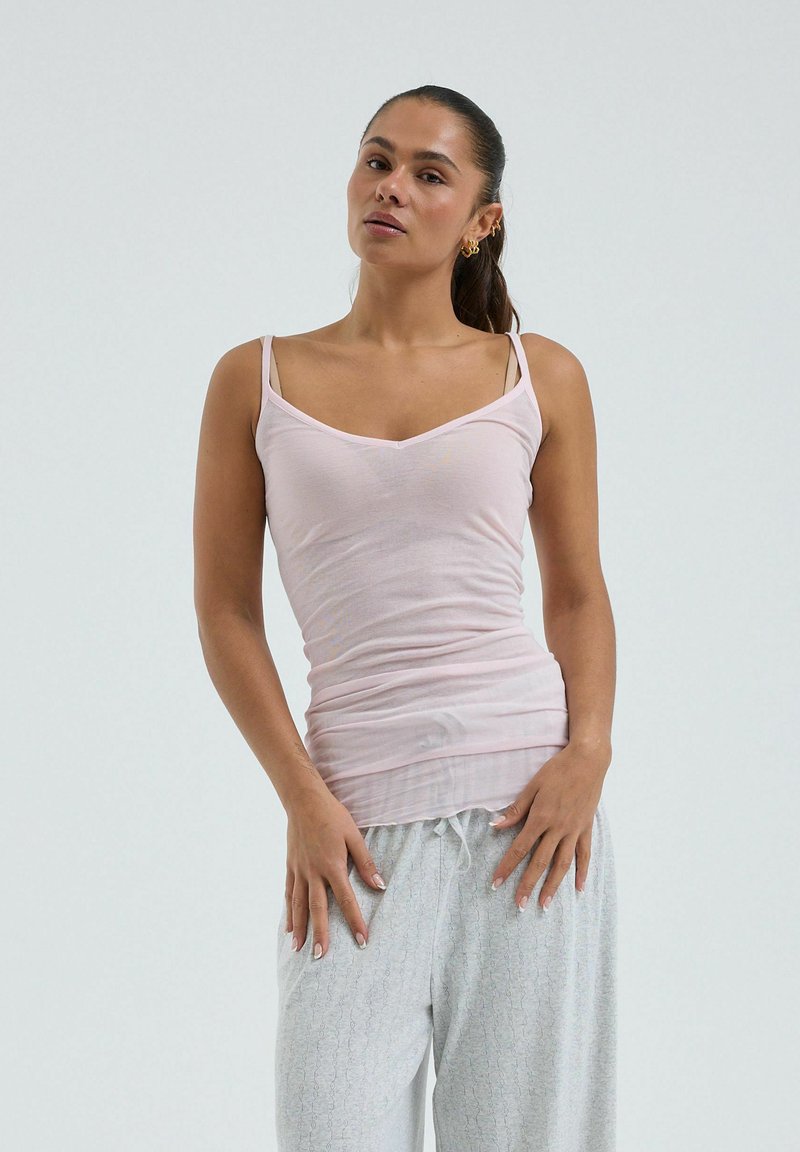 Mujer con cabello oscuro recogido en una coleta, vistiendo una camiseta sin mangas rosa claro y pantalones sueltos gris claro, de pie frente a un fondo liso.