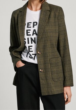 Persoon draagt een groen geruite blazer met twee knopen over een wit grafisch T-shirt en zwarte broek, één hand in de zak, bij kin en knieën bijgesneden.