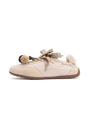 Zapato casual beige con cordones de cinta de satén y dos pompones decorativos unidos cerca de la puntera sobre una suela de goma plana.