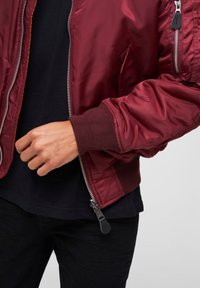 Veste bomber bourgogne avec fermeture éclair, poignets côtelés, poches latérales et matériau synthétique brillant. Portée sur un t-shirt noir.
