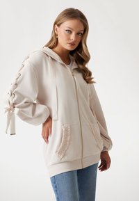 Cream zip-up hoodie with a textured design, oversized sleeves with ribbon ties, and ruffled pocket edges. Soft material and relaxed fit.