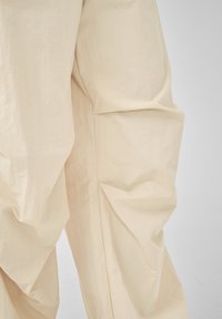 Weit geschnittene beige Hose mit sichtbaren Knitterfalten, dargestellt vom mittleren Oberschenkel bis zum Knöchel vor einem schlichten hellgrauen Hintergrund.