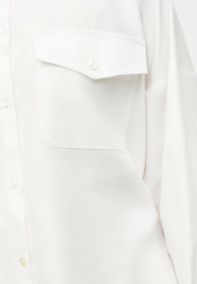 Κοντινό πλάνο σε τσέπη λευκού πουκαμίσου με κουμπωτό καπάκι στην αριστερή πλευρά του στήθους, που δείχνει την υφή του υφάσματος και τα κουμπιά.