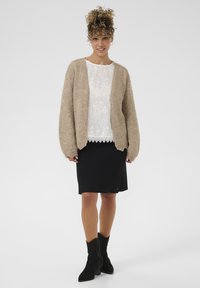 Beige gestrickte Strickjacke über einer weißen Spitzenbluse, kombiniert mit einem knielangen schwarzen Rock und schwarzen Wildleder-Stiefeletten. Sanfte Texturen und weiche Farben.
