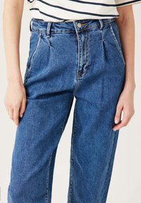 Femme portant un jean bleu loose taille haute et un haut blanc à rayures horizontales noires, se tenant les mains détendues sur les côtés.