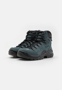 Scarpe da trekking alte in materiale sintetico verde scuro. Presentano lacci neri, dettagli testurizzati e una suola in gomma nera robusta per una buona trazione.