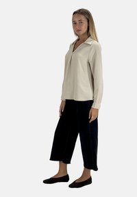 Beiges Langarmshirt mit Kragen und leichter Struktur. Kombiniert mit schwarzen, gekürzten Hosen und flachen Schuhen. Minimalistisches Design.