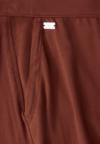 Bruine stof met een gladde textuur, met een zakdetail en een klein wit label dat op de tailleband is genaaid.