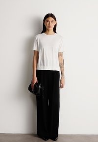 Camiseta blanca de manga corta con cuello redondo, combinada con pantalones sueltos negros. El modelo sostiene un pequeño bolso negro.