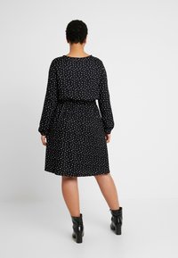 Schwarzes Polka-Dot-Kleid mit langen Ärmeln, tailliertem Schnitt und ausgestelltem Rock. Getragen mit schwarzen Stiefeletten. Glatte Stofftextur.