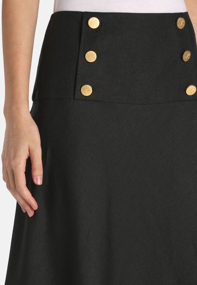 Sort nederdel med høj talje og to lodrette rækker af tre gyldne knapper foran, båret med en lys top og synlig venstre hånd.