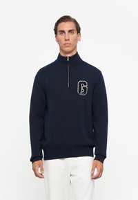 Man wearing a navy blue quarter-zip sweater with a large "G" logo on the chest and white pants against a plain background.