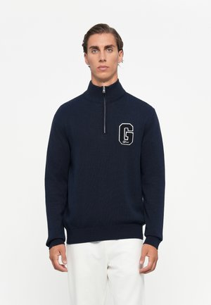 Mann iført en marineblå genser med kvart glidelås og et stort "G"-logo på brystet, samt hvite bukser, mot en ensfarget bakgrunn.