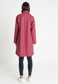 Długi, różowy wodoodporny płaszcz z wysokim kołnierzem i długimi rękawami, wykonany z gładkiego, błyszczącego materiału. Połączony z dopasowanymi dżinsami i białymi trampkami.