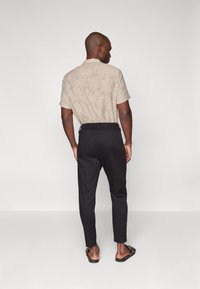 Camisa beige de manga corta con un patrón de hojas, combinada con pantalones negros ajustados y sandalias negras, vista desde la parte de atrás.