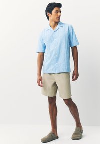 Chemise à manches courtes bleu clair à boutons avec un motif texturé, associée à un short beige et à des sabots gris. Fond mur blanc.