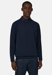 Jeune homme à la peau foncée portant un pull bleu marine par-dessus une chemise à col foncé et un jean bleu, debout devant un fond clair uni.